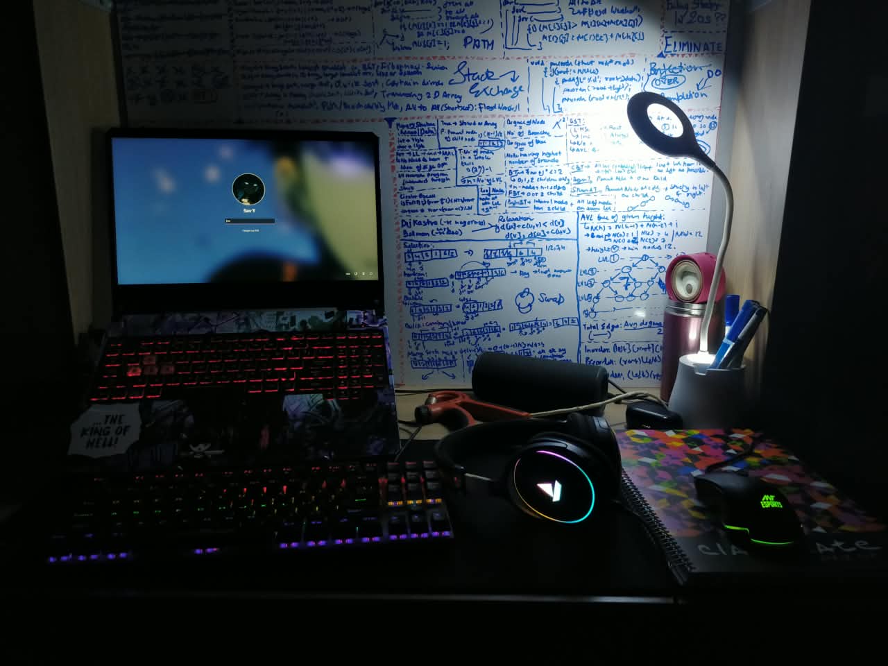 RGB-lit laptop and peripherals beside a wall of handwritten technical notes